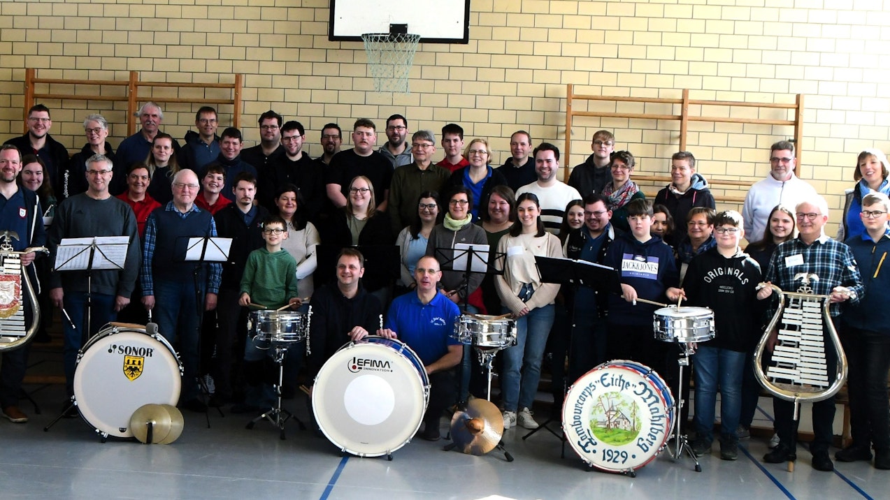 Zahlreiche Musiker stehen in einer Turnhalle. Einige halten Lyras, vor anderen stehen Pauken.