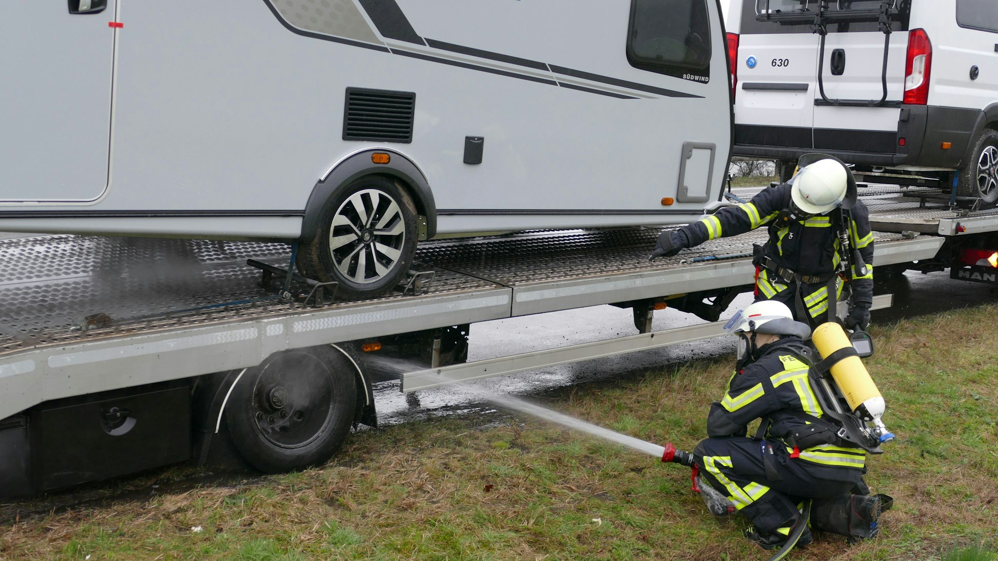 Feuerwehrleute knien neben einem Fahrzeuganhänger. Sie richten den Strahl aus einem Wasserschlauch auf das Fahrgestell.