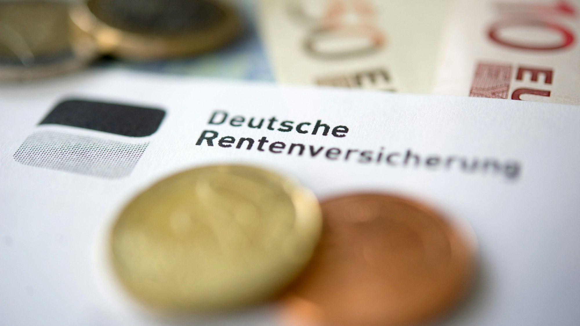 Auf einem Zettel ist das Logo der Deutschen Rentenversicherung zu sehen. Einige Centstücke und Geldscheine liegen auf und unter dem Papier.