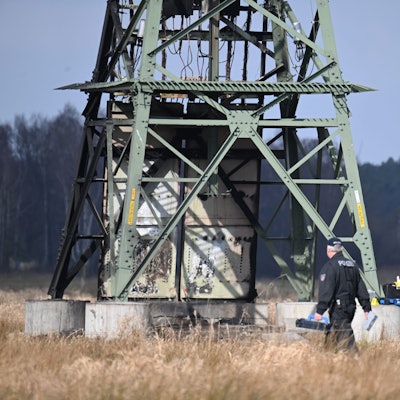 Die Polizei ermittelt an einem beschädigten Strommast auf einem Feld. In der Tesla-Autofabrik in Grünheide steht die Produktion wegen des dadurch verursachten Stromausfalls still.