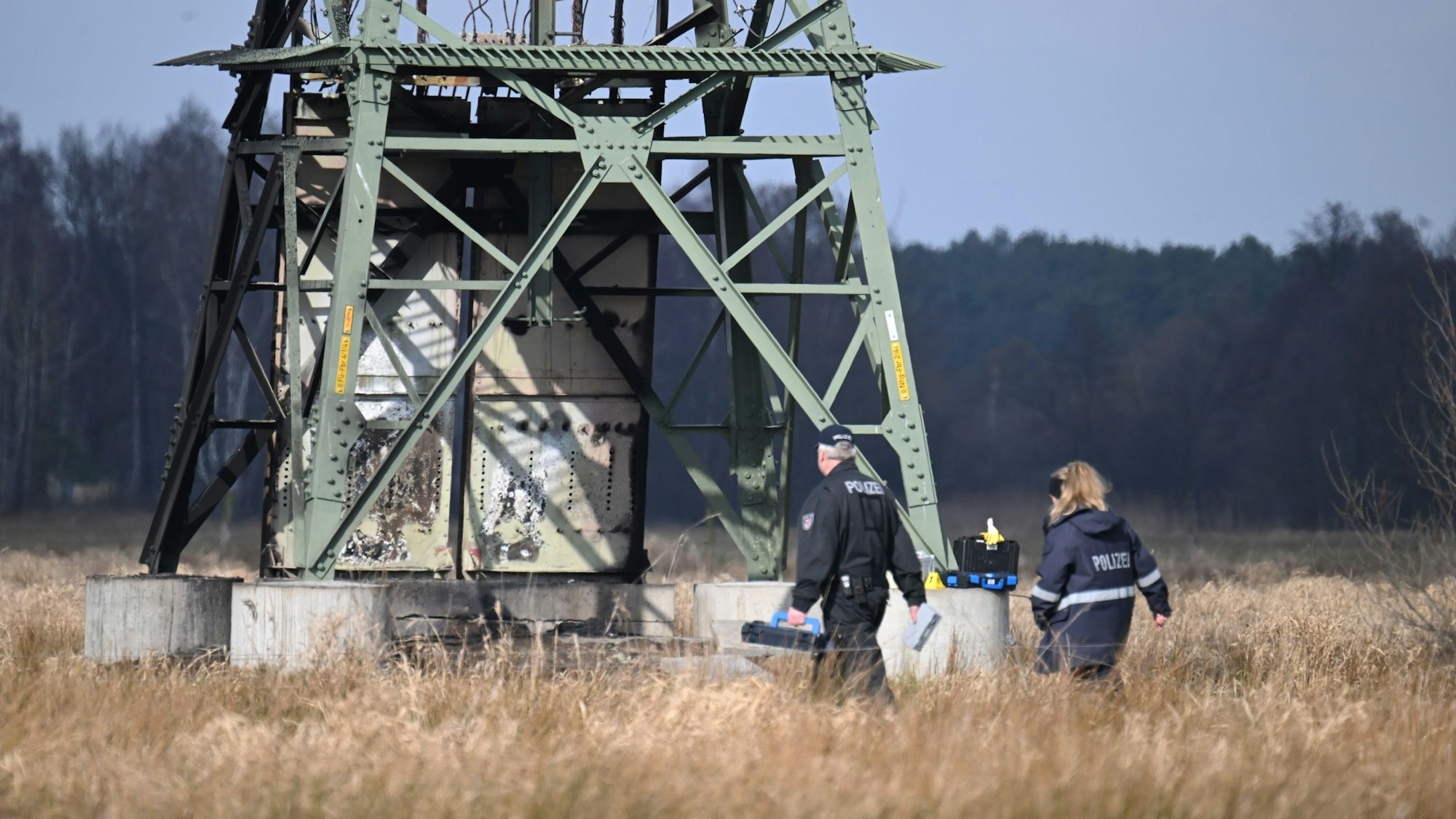 Die Polizei ermittelt an einem beschädigten Strommast auf einem Feld. In der Tesla-Autofabrik in Grünheide steht die Produktion wegen des dadurch verursachten Stromausfalls still.