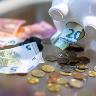 Aus dem Bauch eines Sparschweins kommen Geldscheine und Münzen.