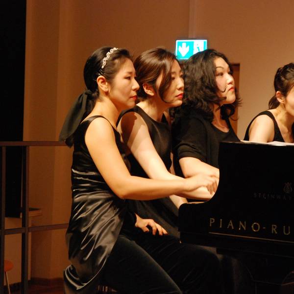 Vier Pianistinnen spielen, nebeneinander sitzend, auf einem Konzertflügel, den Blick konzentriert auf die Noten gerichtet.
