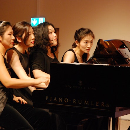 Vier Pianistinnen spielen, nebeneinander sitzend, auf einem Konzertflügel, den Blick konzentriert auf die Noten gerichtet.