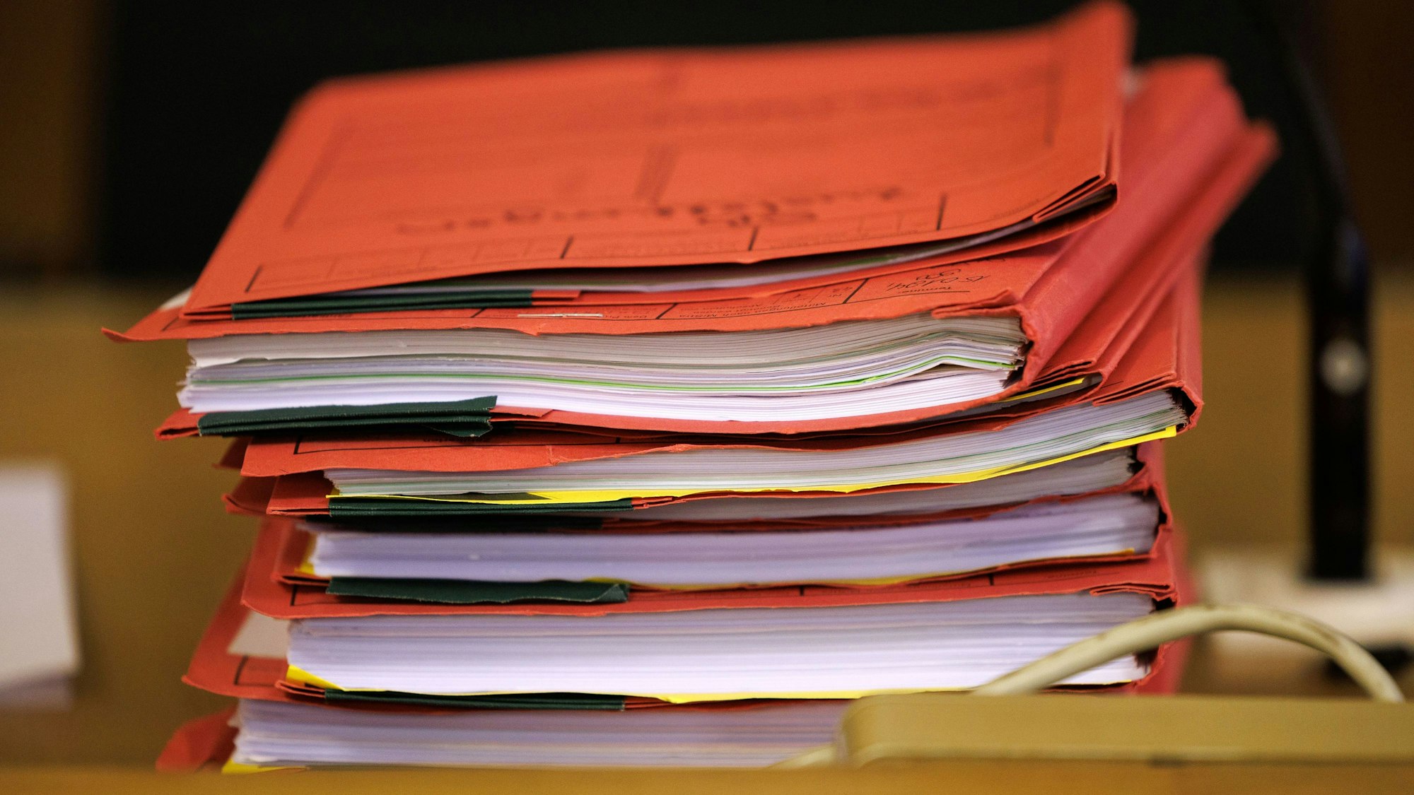 Mehrere rote Prozessakten liegen in einem Gerichtssaal zu Beginn eines Prozesses auf einem Tisch.