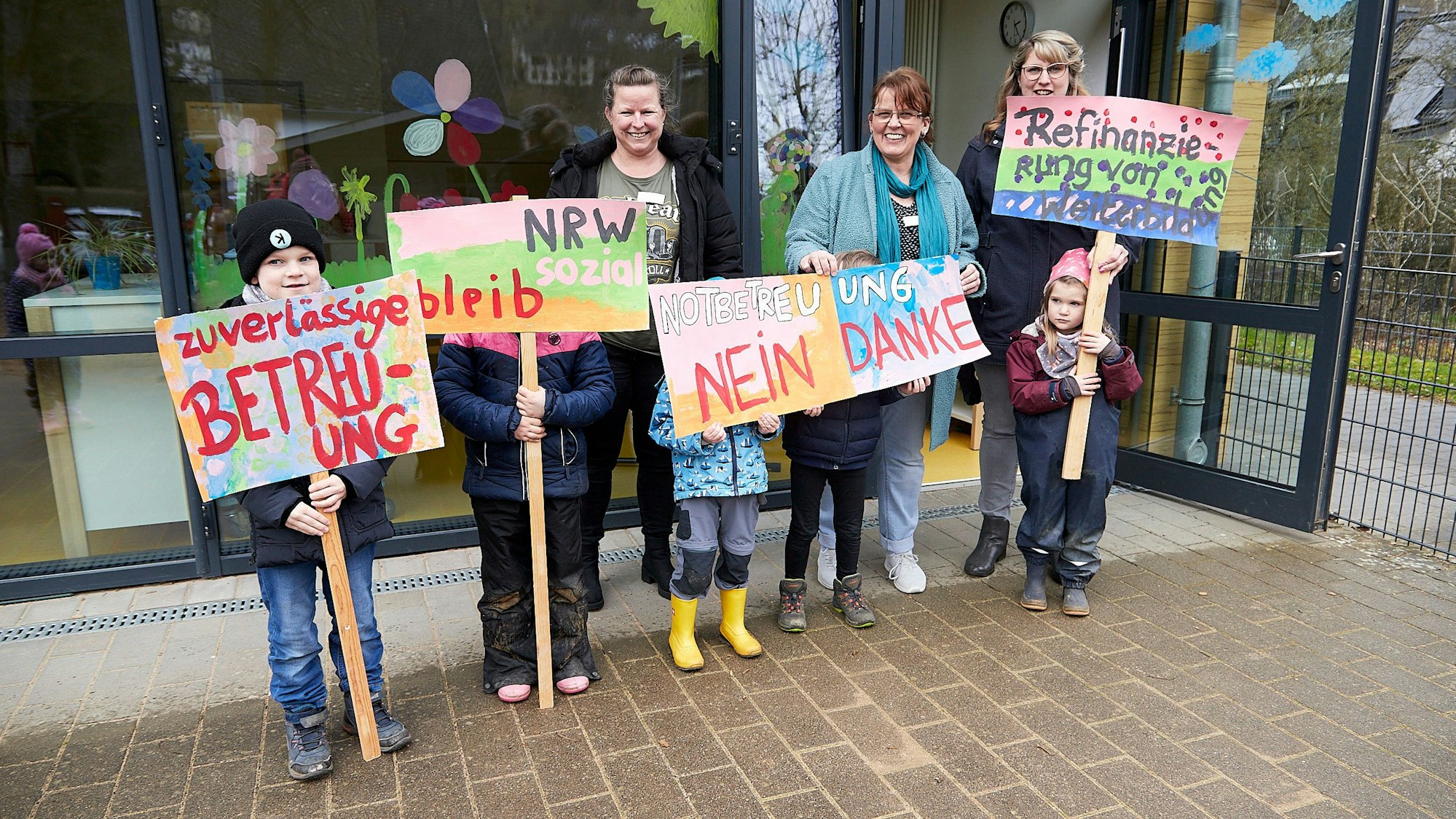 Drei Erzieherinnen und mehrere Kinder stehen mit selbstgemalten Protestplakaten vor der Kita in Schleiden.