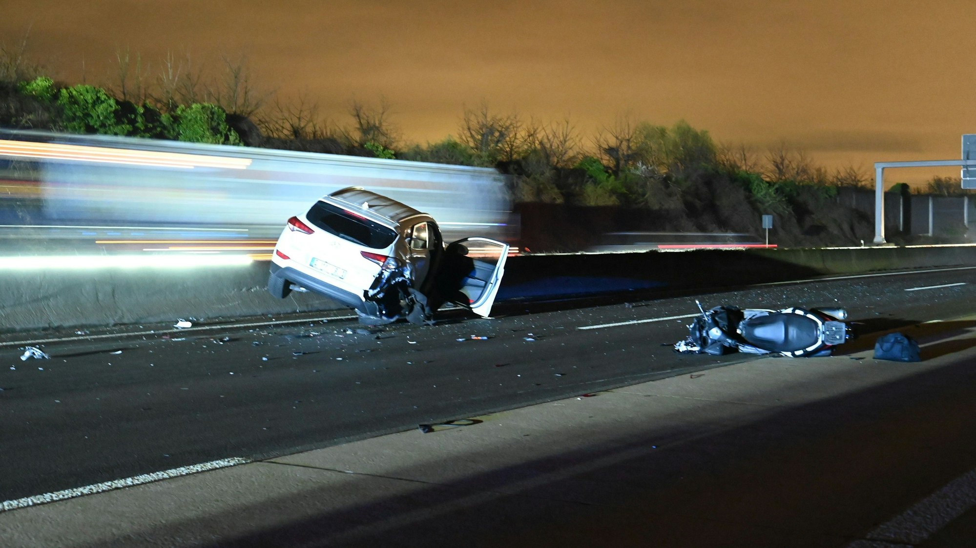Ein Auto liegt schräg auf einer Betonschrammwand, ein Motorrad nicht weit daneben auf der Fahrbahn. Im Hintergrund fahren Lkw vorbei.