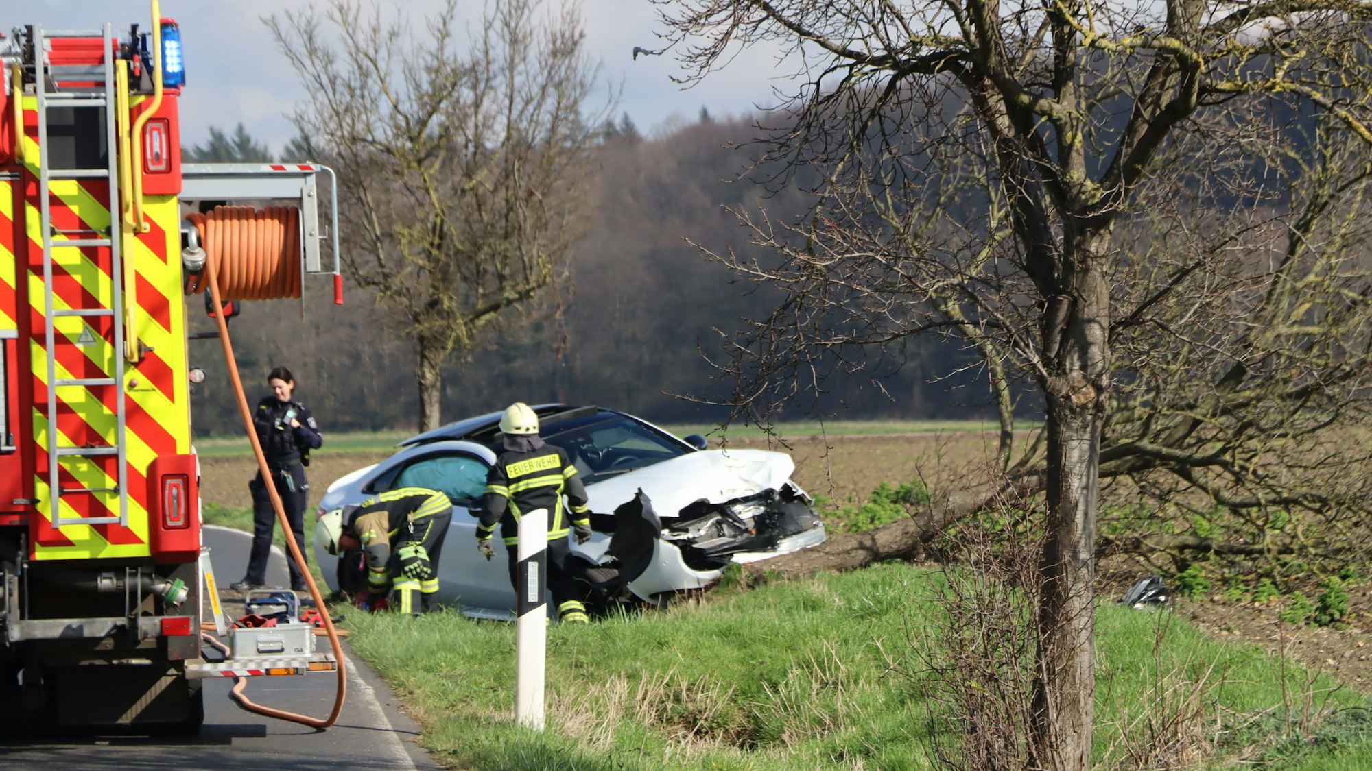 Ein weißes Auto steht zerbeult am Straßenrand. Daneben steht ein Feuerwehrauto und Einsatzkräfte sichern Spuren.Ein umgekippter Baum liegt an der Haube des Autos.