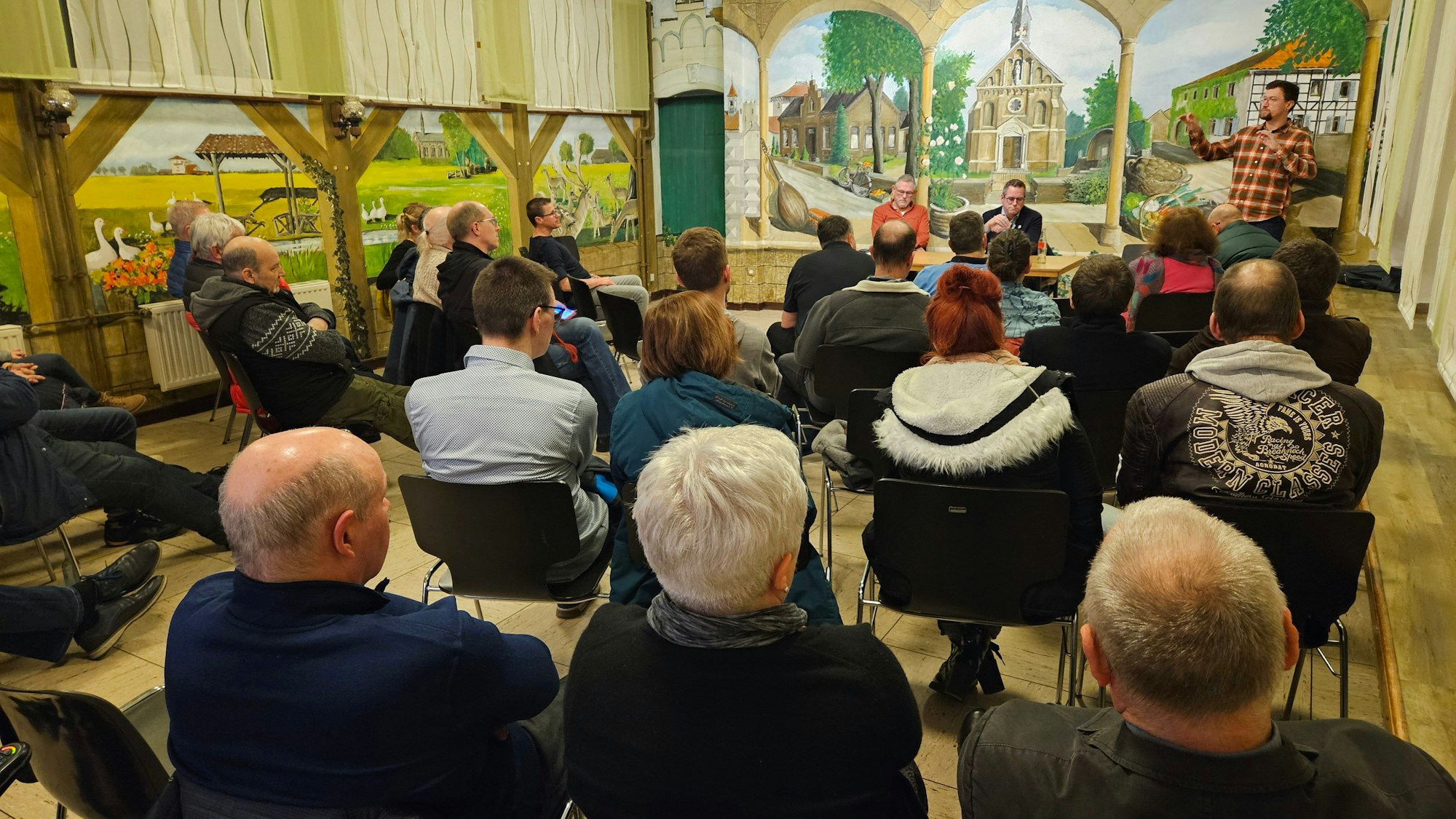 In einem Saal sitzen viele Männer und Frauen in Stuhlreihen, an einem Tisch sitzen zwei Männer, ein Mann steht.