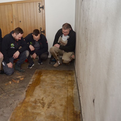 Die vier Männer hocken nebeneinander und schauen sich die Grabplatte an, links die Rückwand des Beichtstuhls.