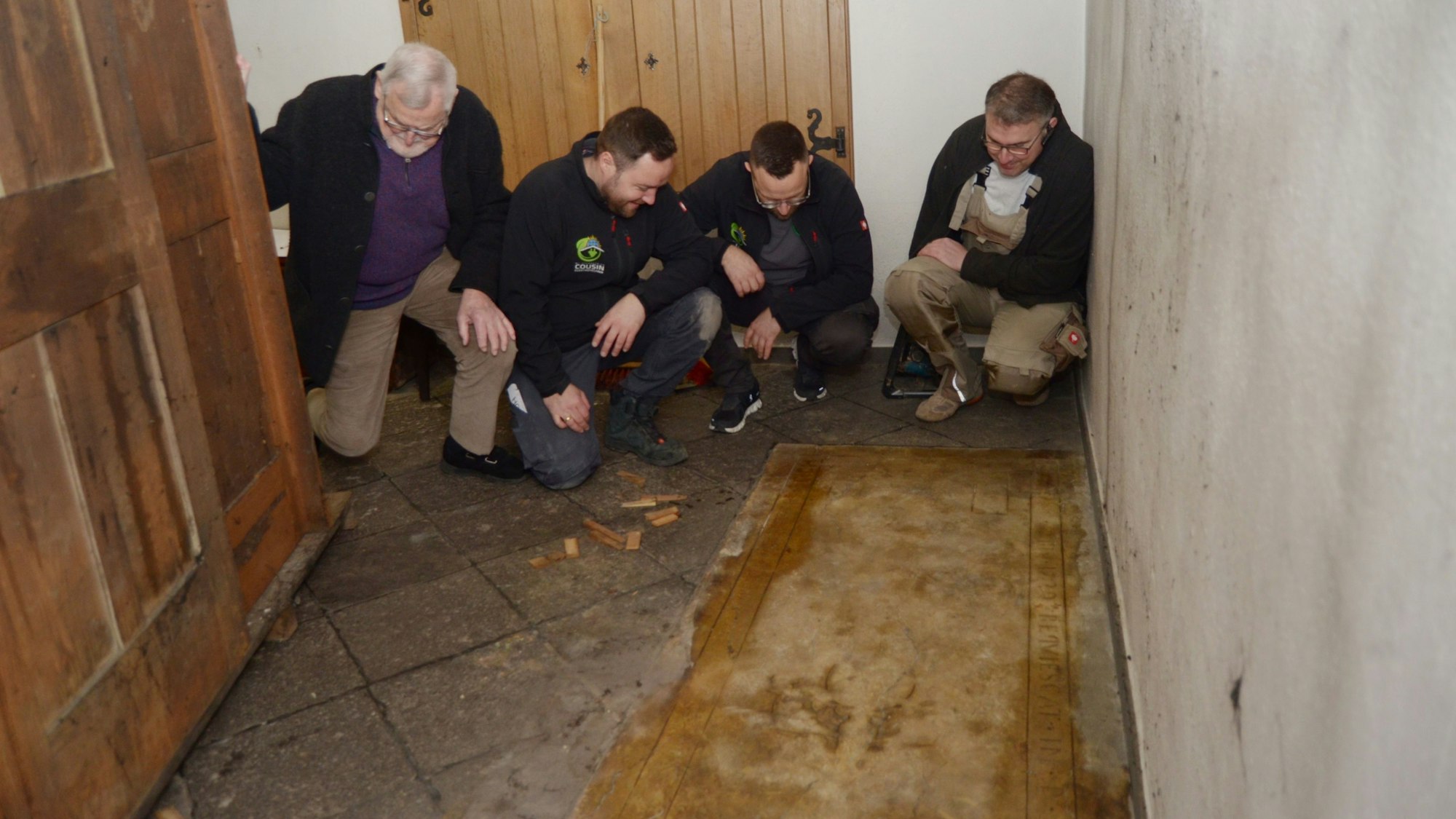 Die vier Männer hocken nebeneinander und schauen sich die Grabplatte an, links die Rückwand des Beichtstuhls.