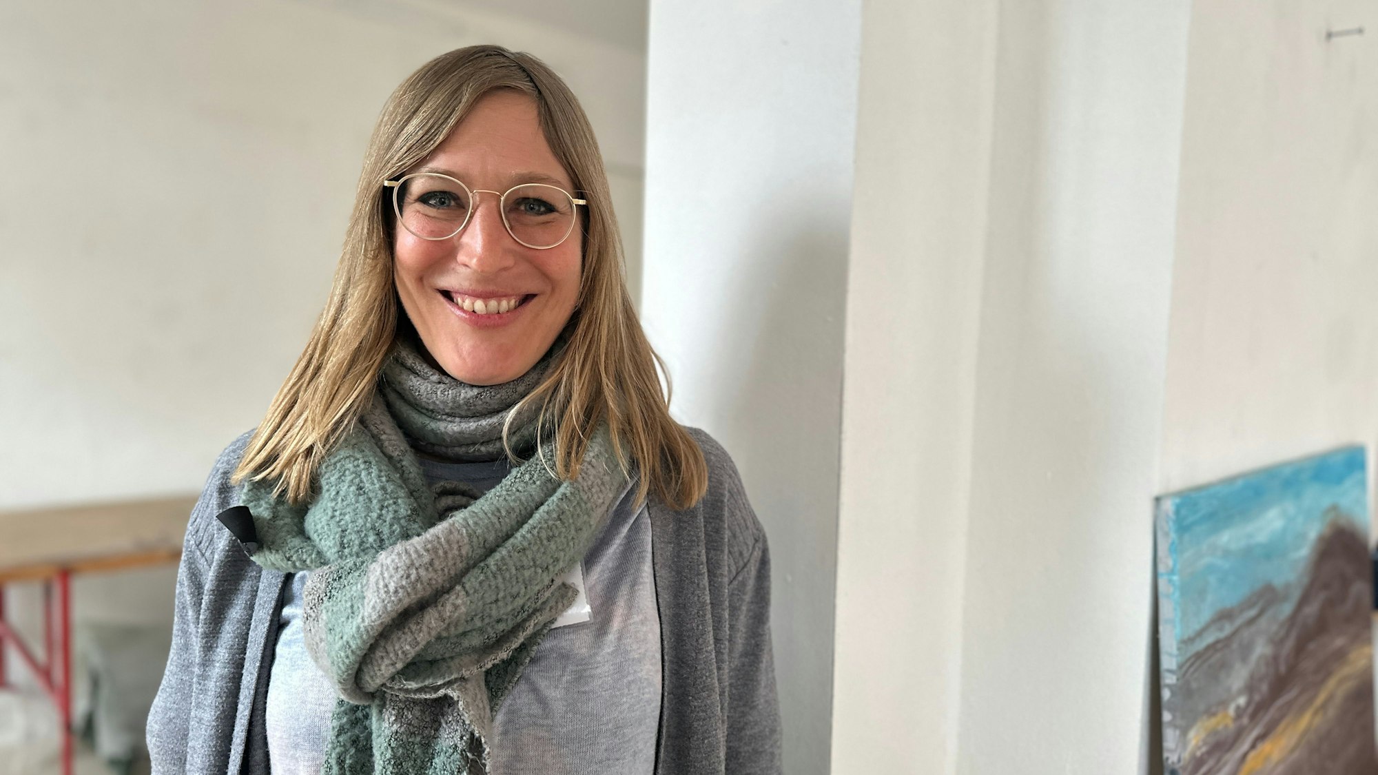 Eine junge Frau mit langem blonden Haar und Brille lacht in die Kamera.