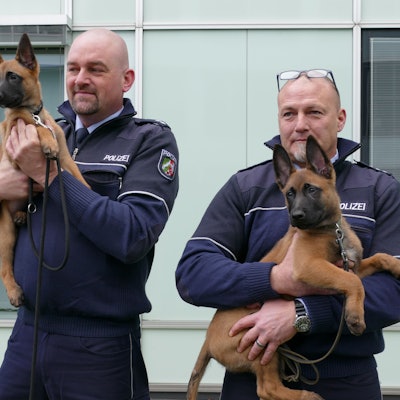 Zwei Polizisten vor einem Wachgebäude. Beide haben jeweils einen Hundewelpen auf dem Arm.