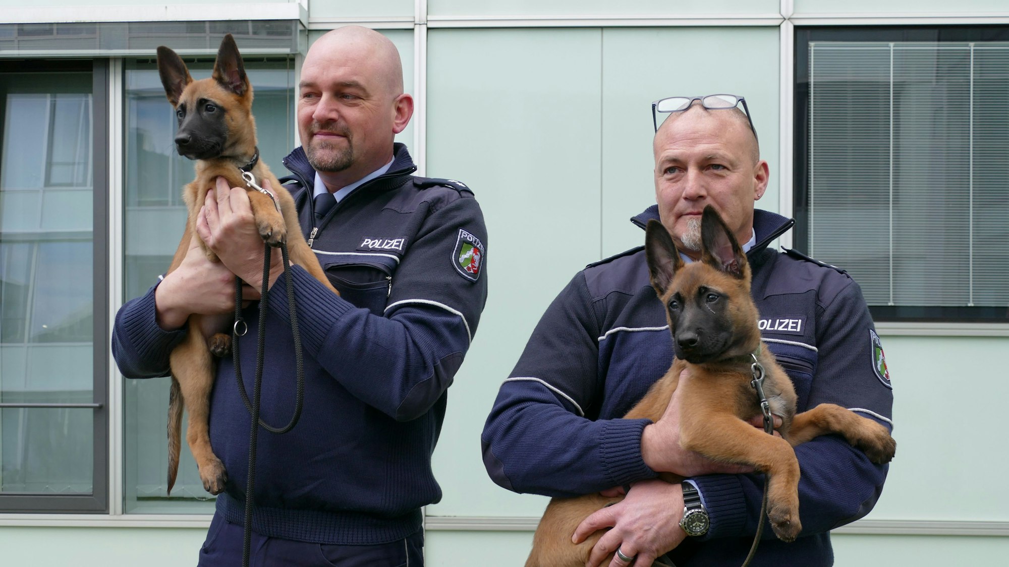 Zwei Polizisten vor einem Wachgebäude. Beide haben jeweils einen Hundewelpen auf dem Arm.
