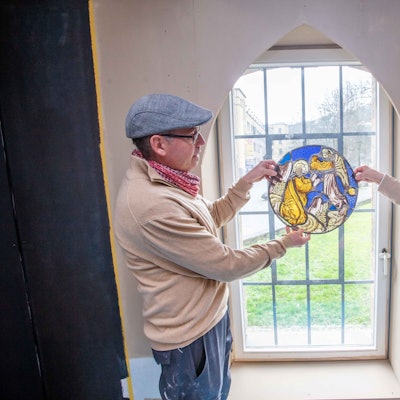 Ein Mann und eine Frau halten ein Glasmosaik in Händen, das Mosaik wird vom Tageslicht erhellt, das durch ein Fenster fällt.
