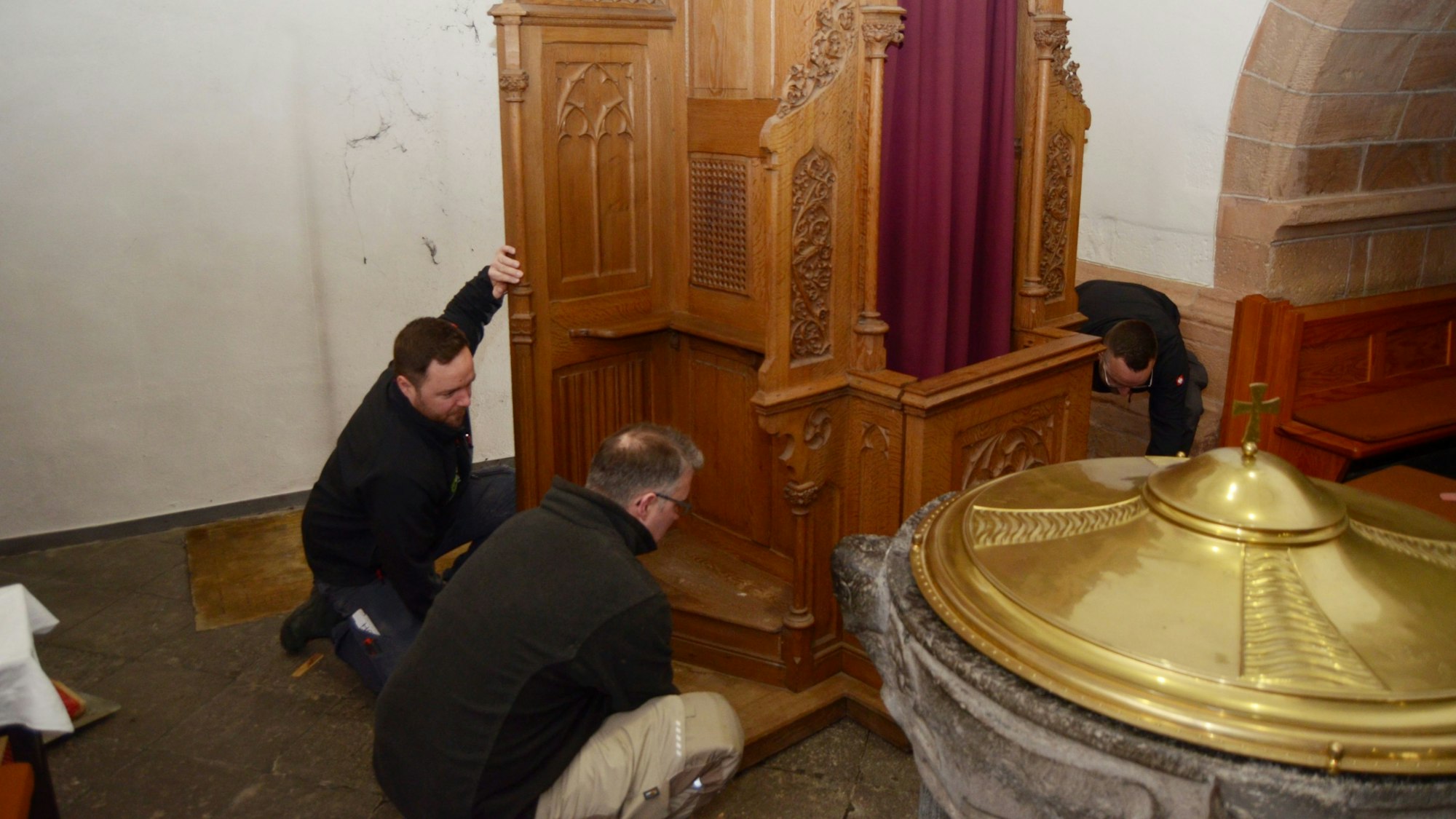 Die drei Männer haben den Beichtstuhl von der Wand weggezogen, im Bildvordergrund steht der Taufstein der Martinskirche.