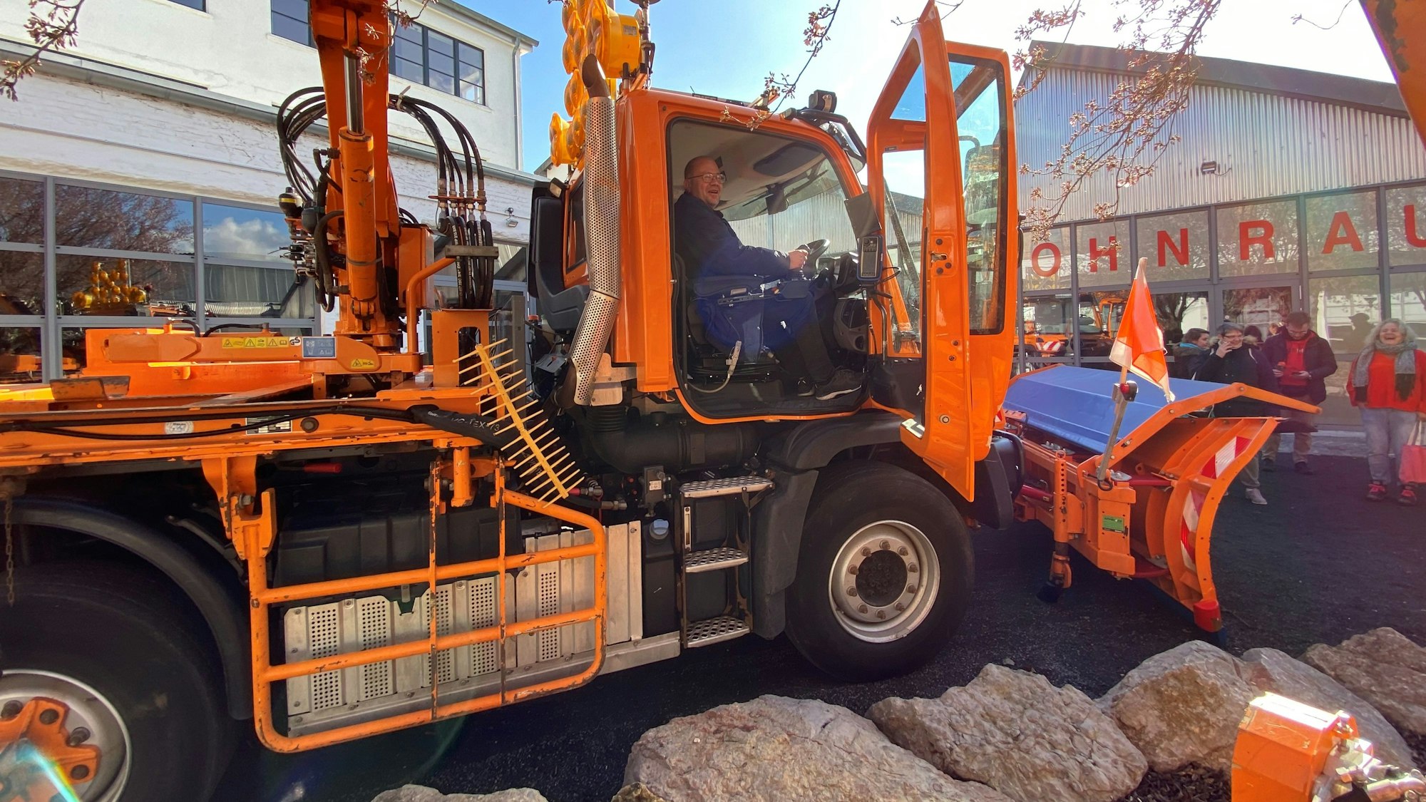 Ein orangefarbenes Fahrzeug des Winterdienstes mit einem Räumschild steht auf einem Parkplatz. Ein Mann sitzt am Steuer, einige Besucher einer Berufswahlmesse schauen sich das Fahrzeug an.