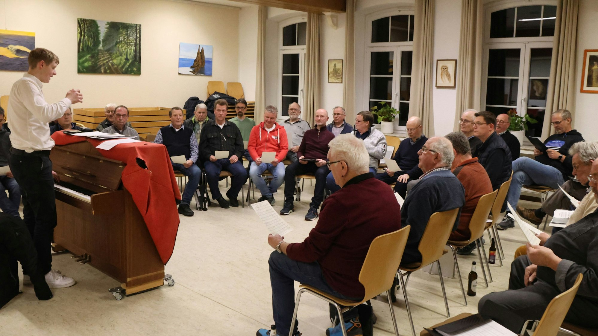 Linus Stahl steht in einem weißen Hemd am Klavier und dirigiert die vor ihm sitzende Chorgemeinschaft MGV Harmonie Wendershagen in einem Proberaum.