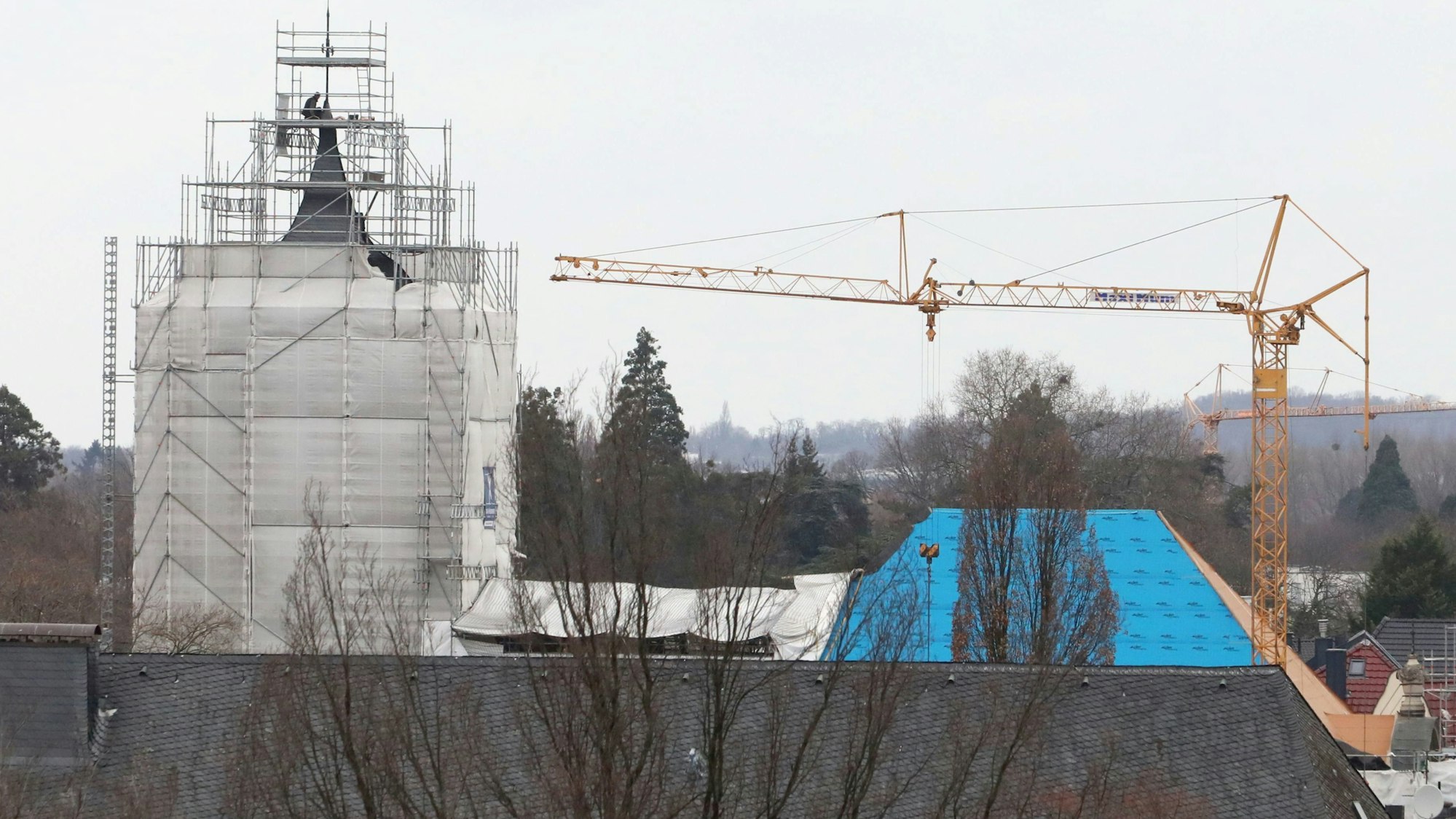 Der Turm einer Kirche ist eingerüstet und mit Planen verhangen, neben der Kirche steht ein gelber Baukran.