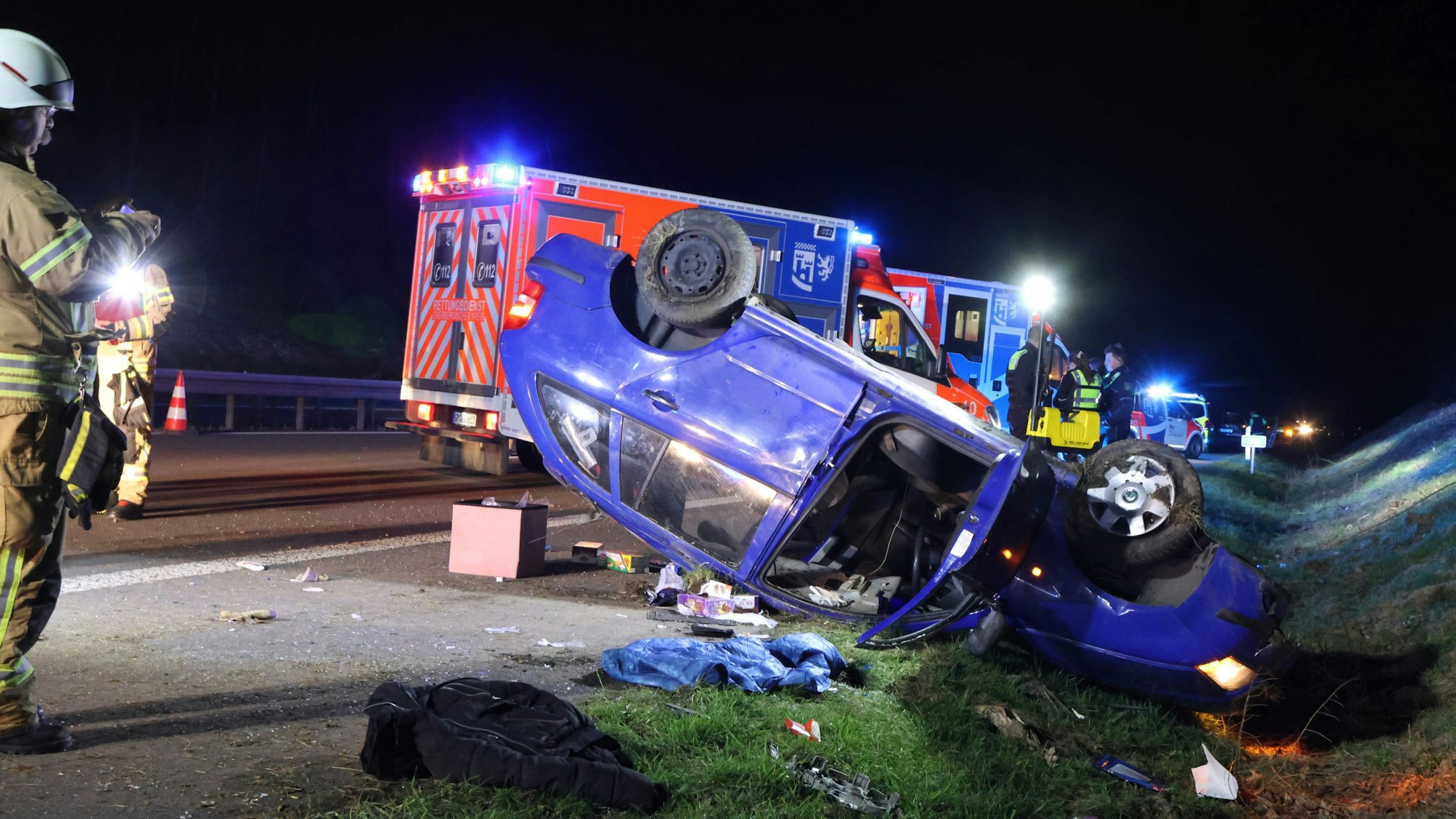 Ein Unfallwagen liegt am Rand einer Autobahn auf dem Dach. Ein Rettungswagen ist im Hintergrund zu sehen. Vorne steht ein Feuerwehrmann.