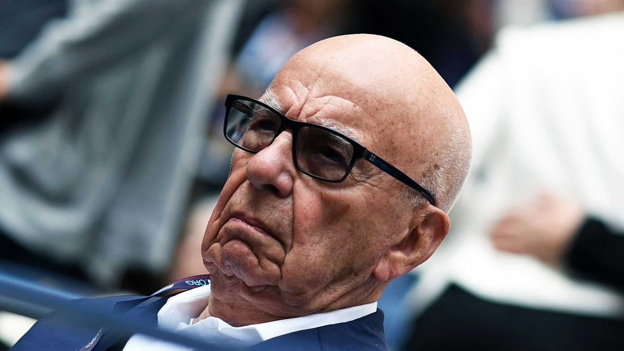 Rupert Murdoch sitzt neben unscharf gestellten Personen und trägt eine Sonnenbrille.