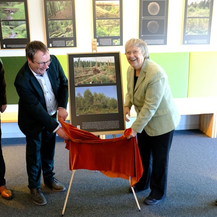 Eröffnung der Sonderausstellung Wilde Bilder. Oliver Krischer und Bärbel Höhn stehen vor einer Staffelei, auf der zwei Fotos präsentiert werden, sie halten ein rotes Tuch. Daneben steht Michael Lammertz mit einem Mikrofon.