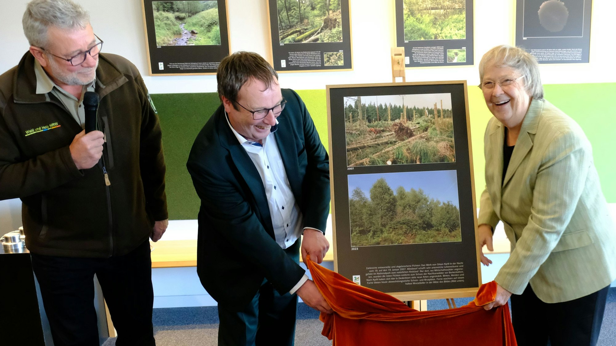 Eröffnung der Sonderausstellung Wilde Bilder. Oliver Krischer und Bärbel Höhn stehen vor einer Staffelei, auf der zwei Fotos präsentiert werden, sie halten ein rotes Tuch. Daneben steht Michael Lammertz mit einem Mikrofon.