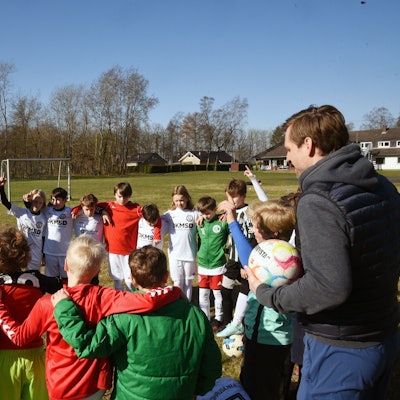 Eine Gruppe Kinder und zwei Trainer stehen in einem Kreis auf einem Fußballplatz zusammen.