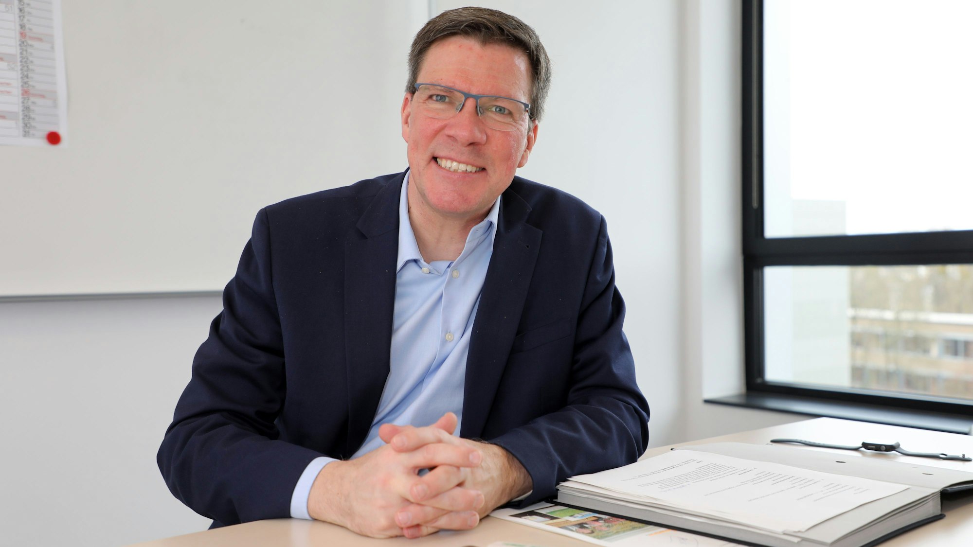 Landrat Stephan Santelmann sitzt an einem Tisch im Kreishaus am Rübezahlwald in Bergisch Gladbach und lächelt in die Kamera.