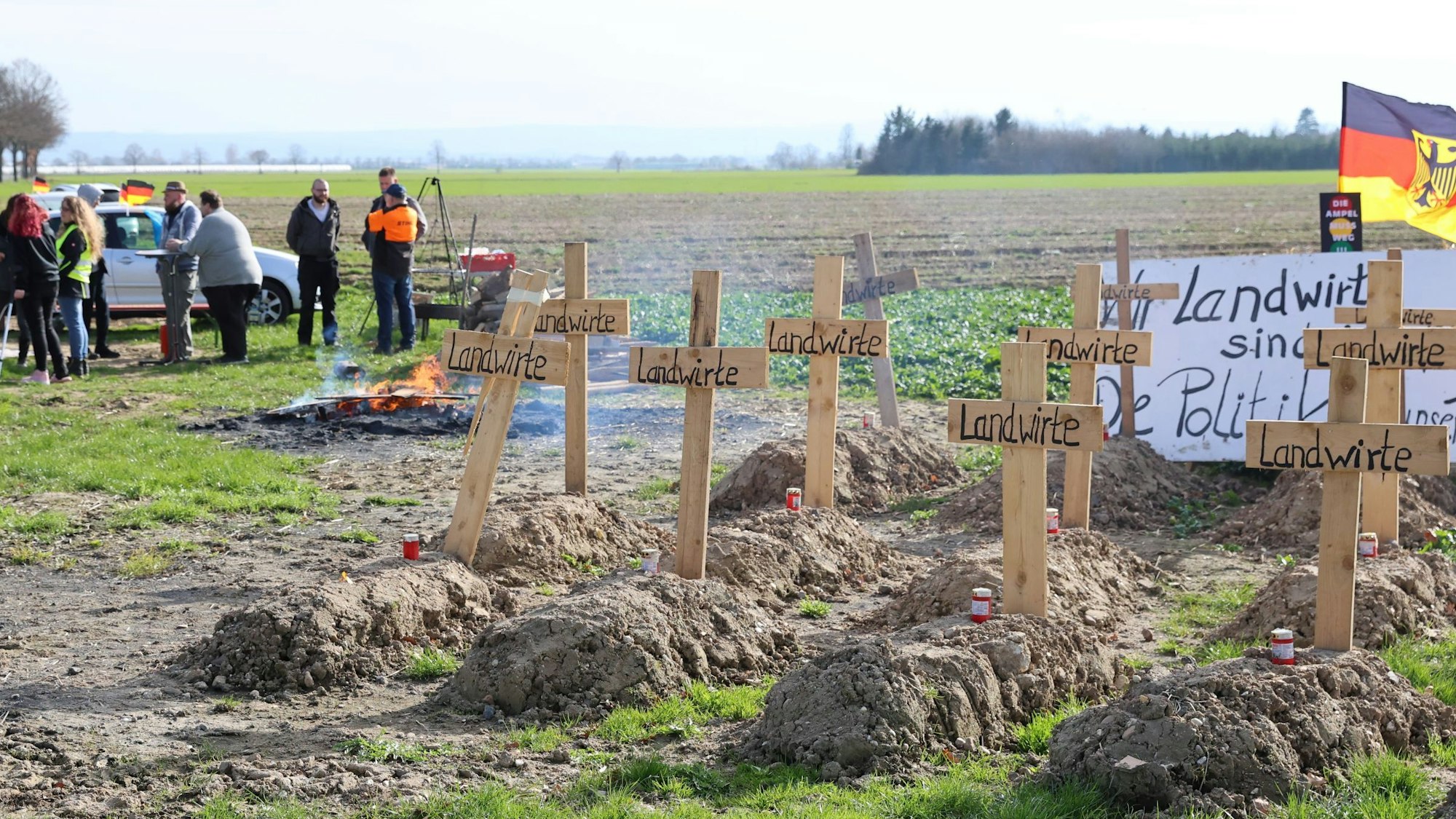Eine Gruppe Menschen steht an einem Mahnfeuer auf einem Feld. Im Vordergrund sind symbolische Gräber zu sehen, die im Rahmen der Bauernproteste angelegt wurden.