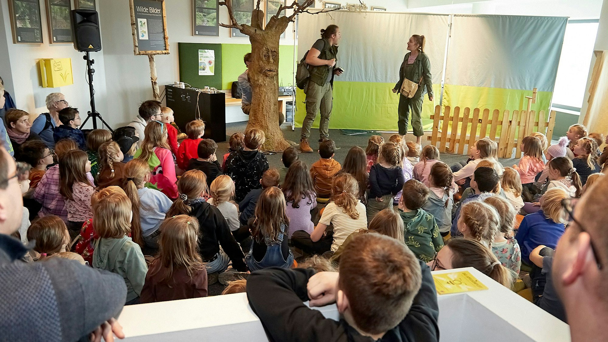Zahlreiche Kinder sind von hinten zu sehen. Sie sitzen in einem Raum auf dem Boden und schauen der Aufführung des Theaterstücks „Das Apfelkompott“ zu.