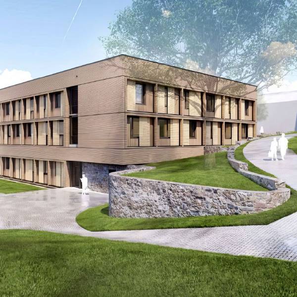 Ein langgestreckter Bau aus Holz ist umgeben von Fußwegen und einer Grünanlage.