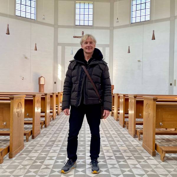 Ein blonder Mann mit Daunenjacke und Umhängetasche steht in einem hellen, großen Raum mit Kirchenbänken und gemustertem Fußboden.