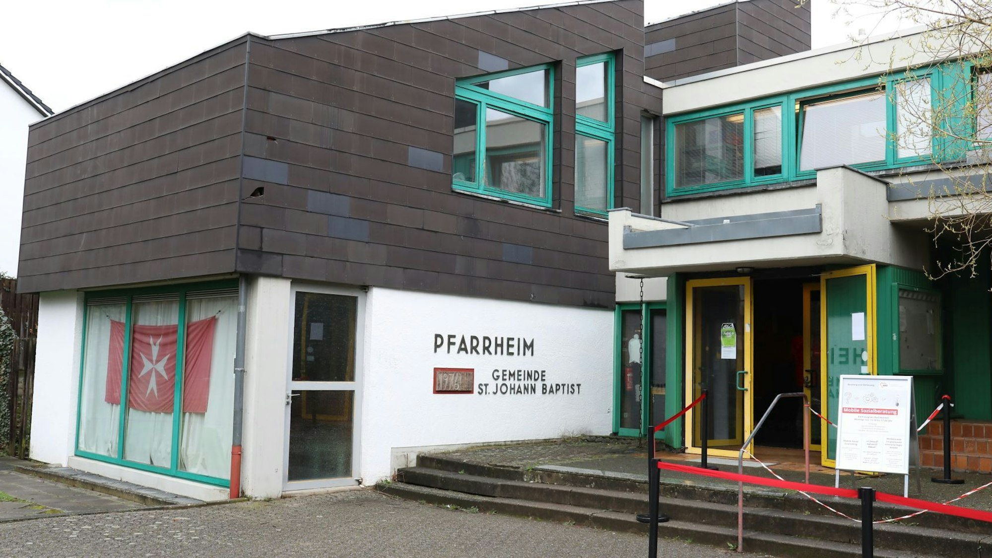 Ein Gebäude mit dem Schriftzug Pfarrheim St. Johann Baptist an der Fassade. Die Tür des Hauses steht offen.