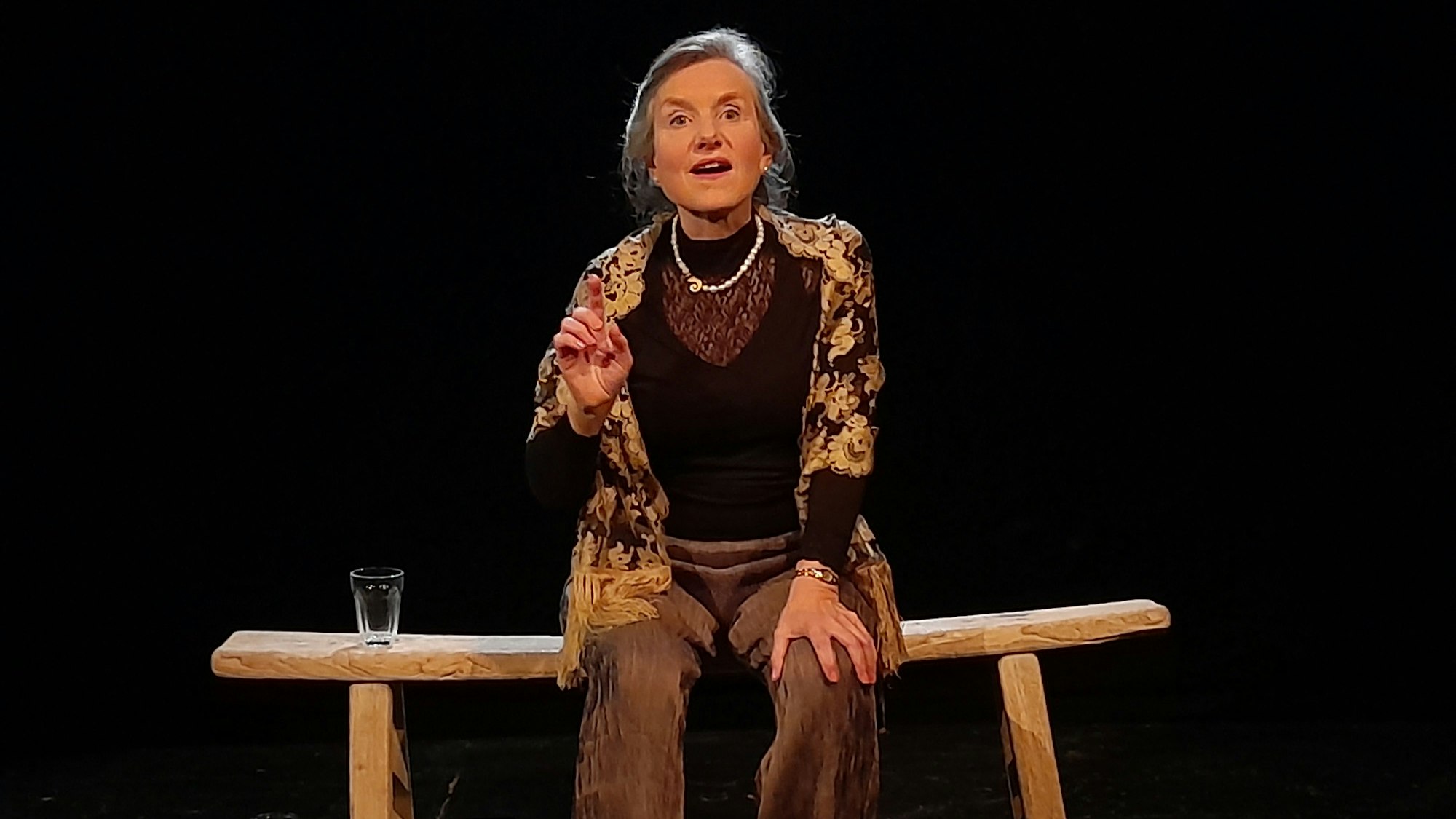 Eine ältere Frau mit Zopf sitzt auf einer Bank. Sie spricht mit erhobener Hand.