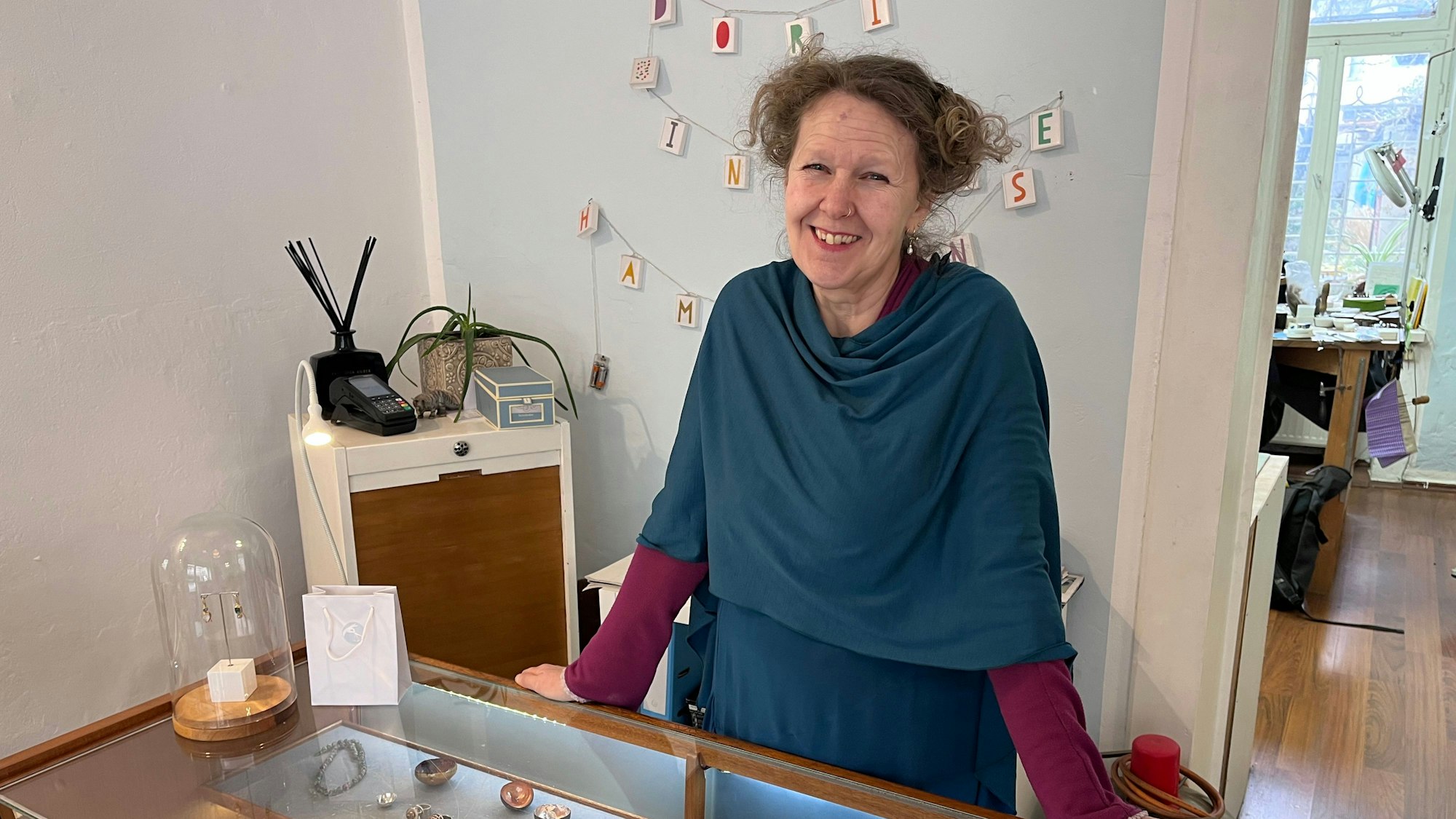 Eine Frau mit hochgestecktem Haar steht lachend hinter einer Juwelier-Theke.