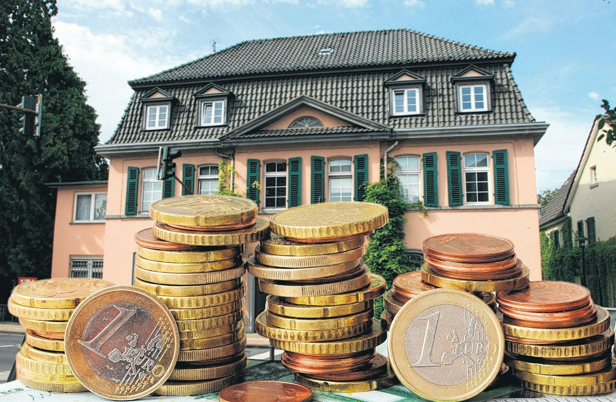 Ein rosafarbenes Haus mit grünen Fensterläden im Hintergrund, vorne einige hohe Stapel Euro-Münzen.