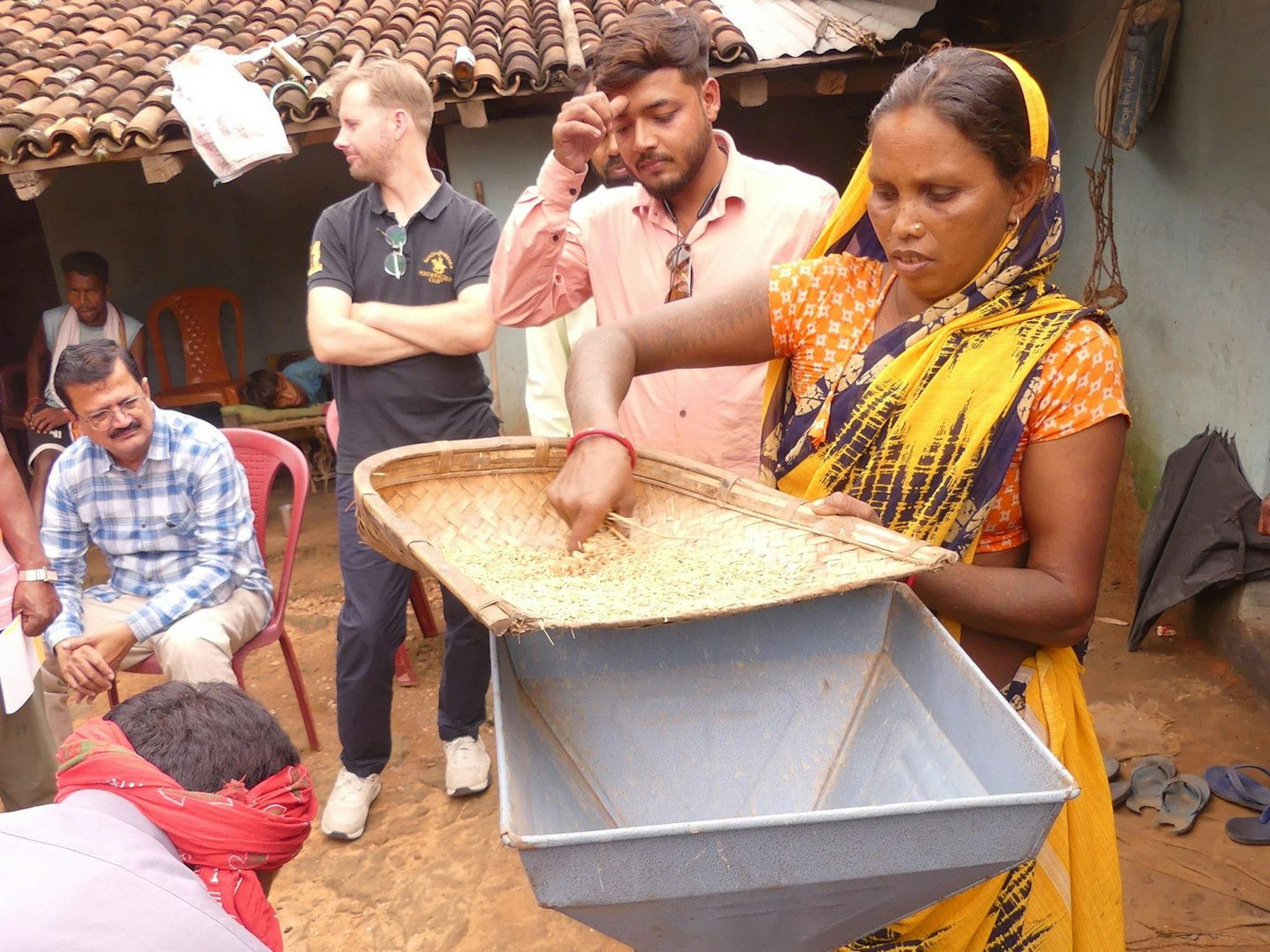 Eine Frau, die einen traditionellen Sari trägt, füllt Reis in den Trichter einer Mühle. Mehrere Menschen stehen um sie herum.