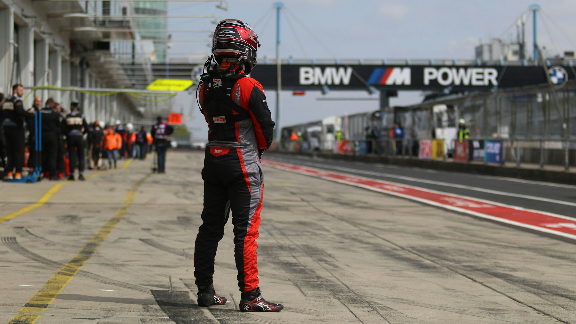 Rennfahrer Alex Kroker steht in voller Montur mit dem Rücken zur Kamera in der fast leeren Boxengasse.