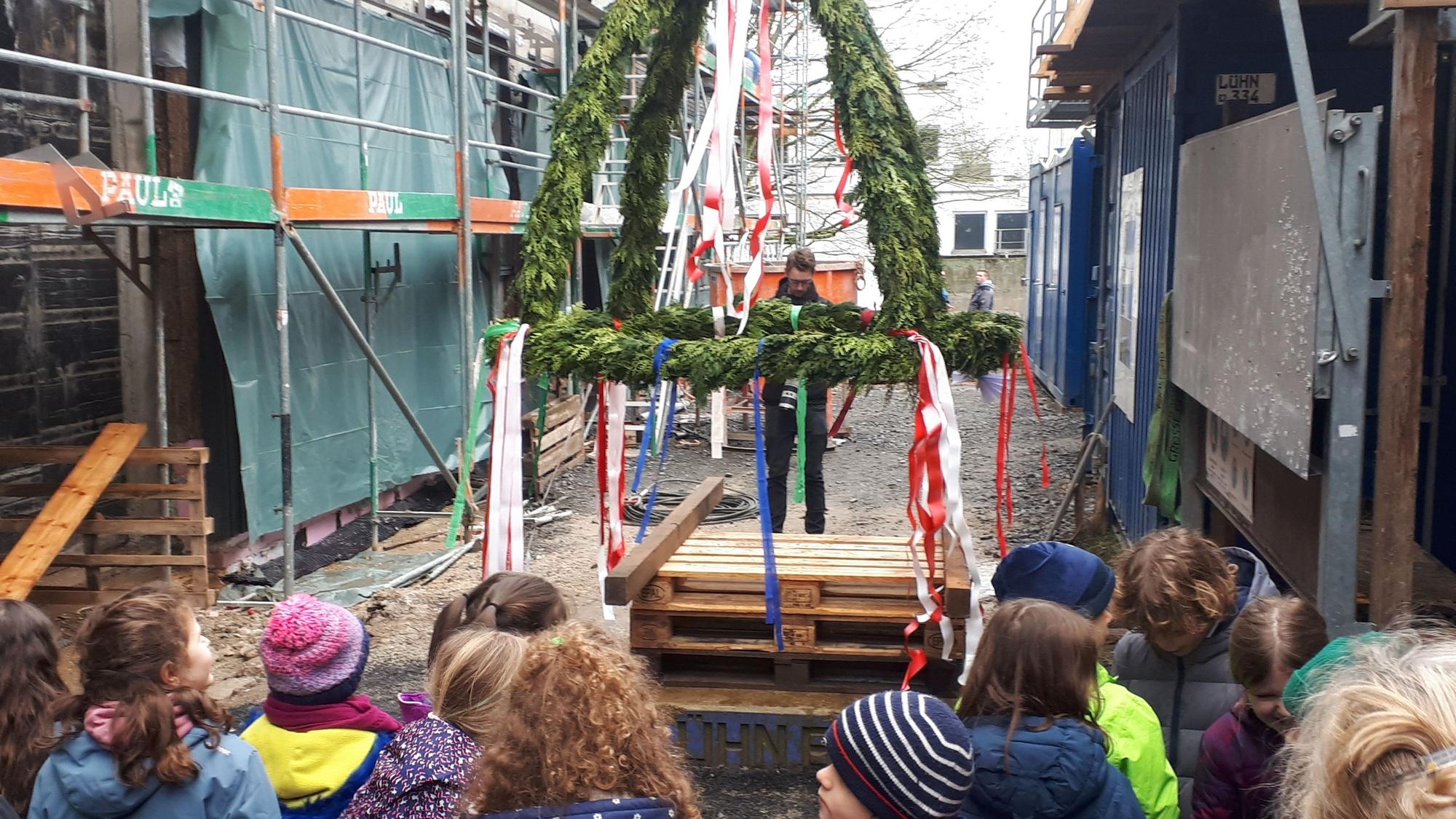 Kinder stehen auf einer Baustelle vor einem festlich geschmückten Richtkranz.