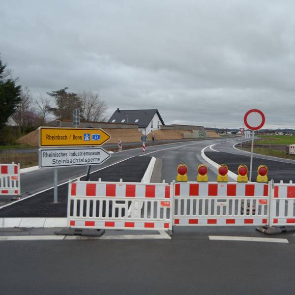 Die Zufahrt in die neu gebaute Umgehungsstraße am Ortsrand von Kuchenheim ist noch durch rot-weiße Sperrzäune blockiert. Auf einer Verkehrsinsel stehen Schilder, die in Richtung Rheinbach, Bonn, zur Steinbachtalsperre und zum Industriemuseum weisen.