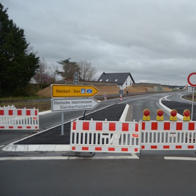 Die Zufahrt in die neu gebaute Umgehungsstraße am Ortsrand von Kuchenheim ist noch durch rot-weiße Sperrzäune blockiert. Auf einer Verkehrsinsel stehen Schilder, die in Richtung Rheinbach, Bonn, zur Steinbachtalsperre und zum Industriemuseum weisen.