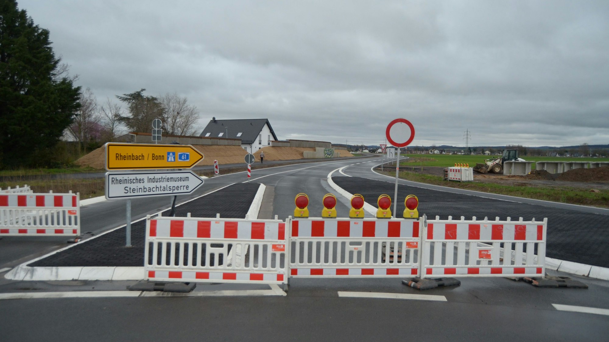 Die Zufahrt in die neu gebaute Umgehungsstraße am Ortsrand von Kuchenheim ist noch durch rot-weiße Sperrzäune blockiert. Auf einer Verkehrsinsel stehen Schilder, die in Richtung Rheinbach, Bonn, zur Steinbachtalsperre und zum Industriemuseum weisen.