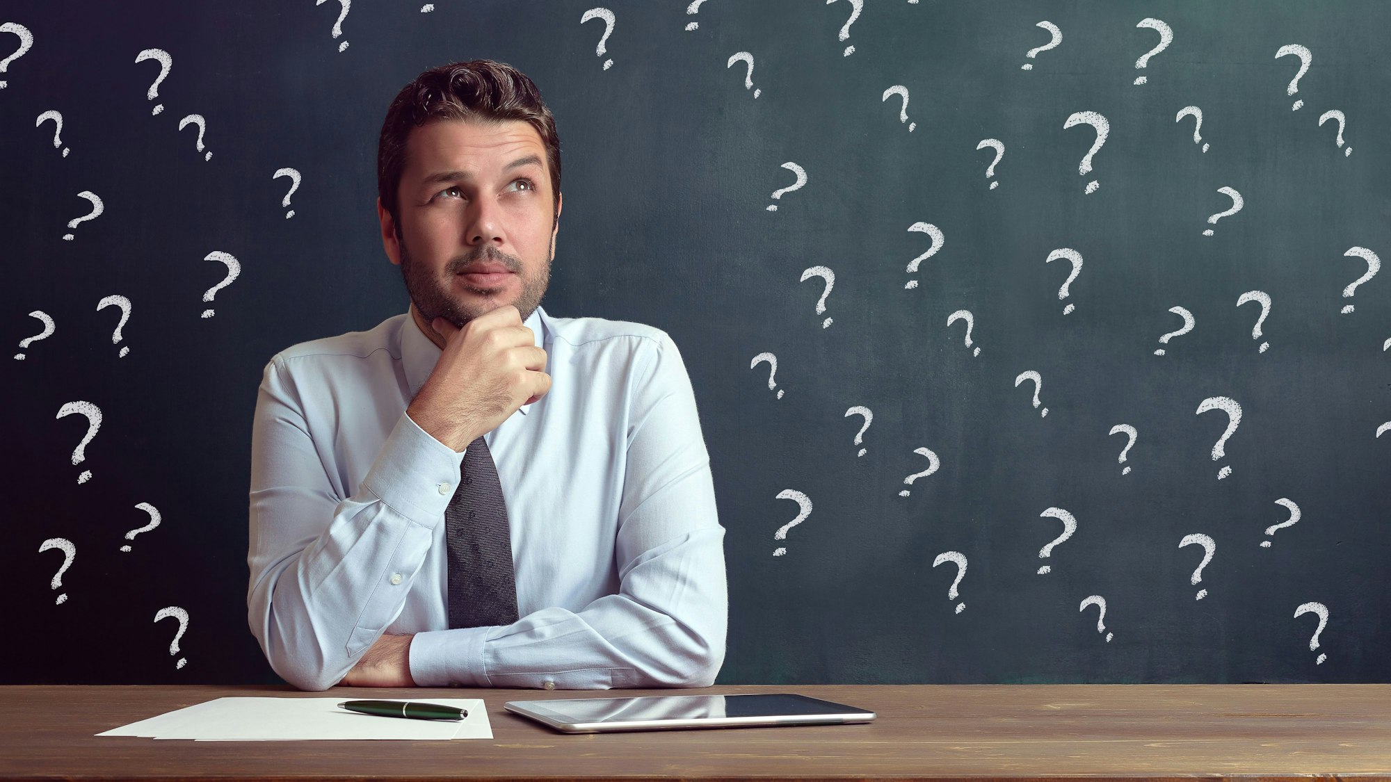 Mann sitzt am Schreibtisch, hinter ihm viele Fragezeichen.