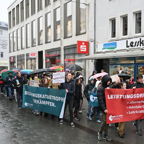 Vorneweg gehen Schüler und tragen ein Banner. Es folgen weitere Demonstranten, teils mit Plakaten, teils mit Regenschirmen in der Hand. Im Hintergrund sieht man die Fassaden von Geschäften.