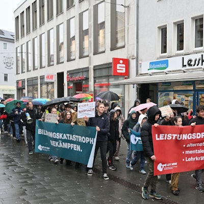 Vorneweg gehen Schüler und tragen ein Banner. Es folgen weitere Demonstranten, teils mit Plakaten, teils mit Regenschirmen in der Hand. Im Hintergrund sieht man die Fassaden von Geschäften.
