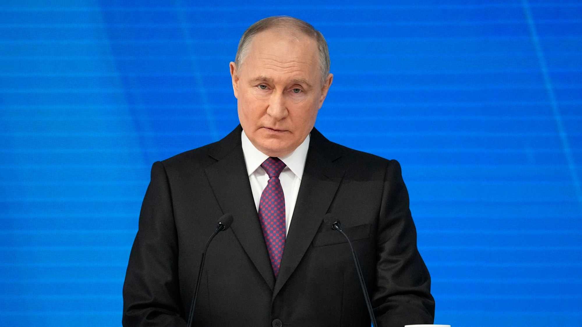 Der russische Präsident Wladimir Putin steht bei einer Rede vor einem Pult auf einer Bühne. Mit seinem schwarzen Anzug steht er vor einer blau angeleuchteten Wand.