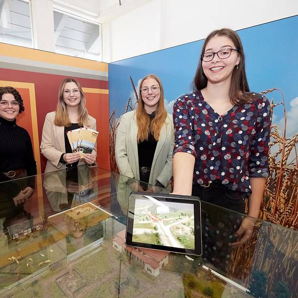 Vier junge Frauen stehen in einem Ausstellungsraum an einem Glastisch, eine hält ein Tablet in der Hand.