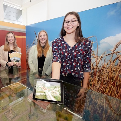 Vier junge Frauen stehen in einem Ausstellungsraum an einem Glastisch, eine hält ein Tablet in der Hand.