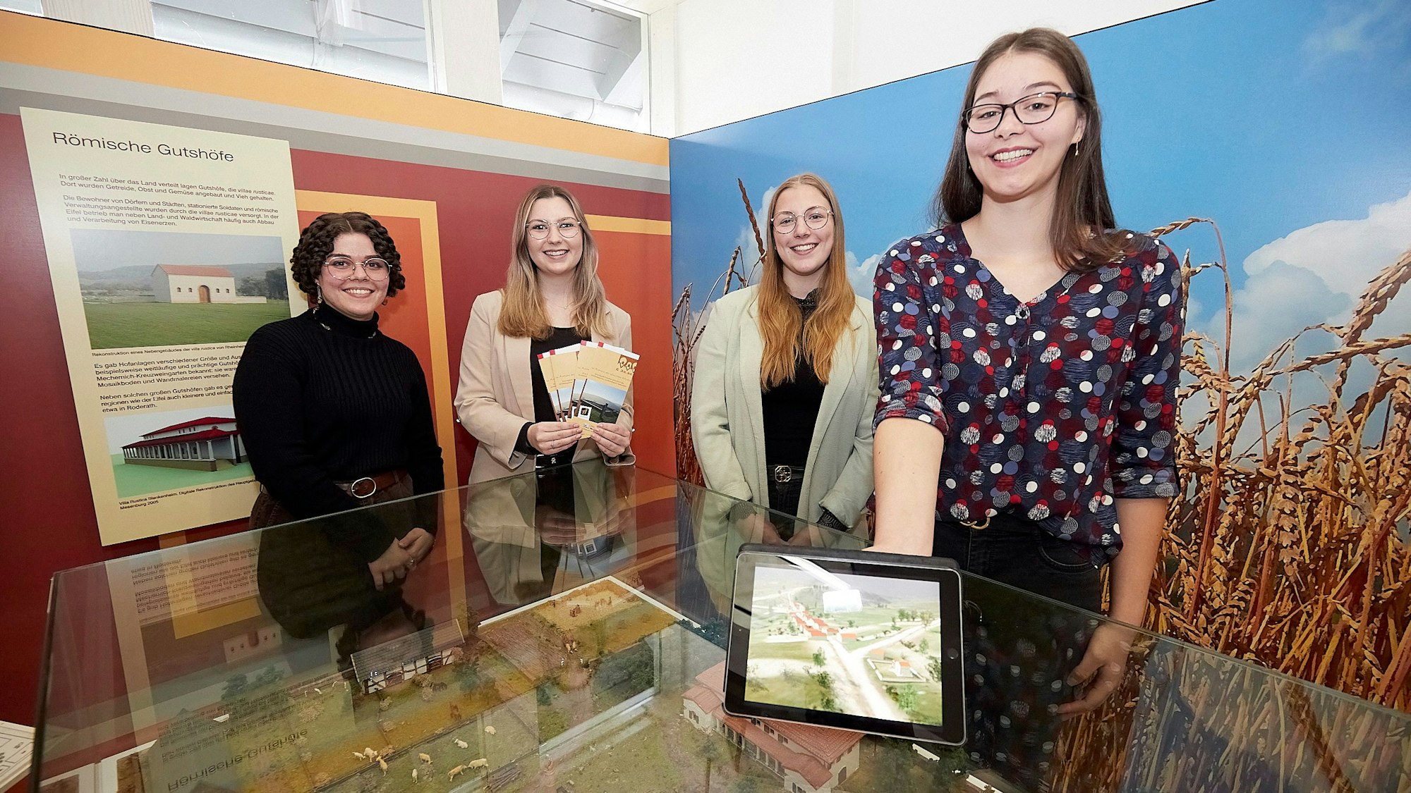 Vier junge Frauen stehen in einem Ausstellungsraum an einem Glastisch, eine hält ein Tablet in der Hand.